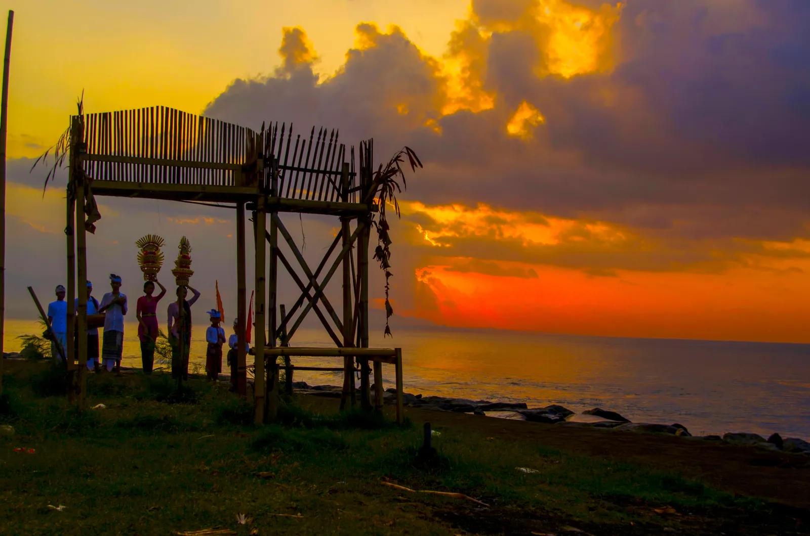 Beberapa orang dan struktur bambu di tepian Pantai Keramas dengan laut dan langit berwarna keemasan