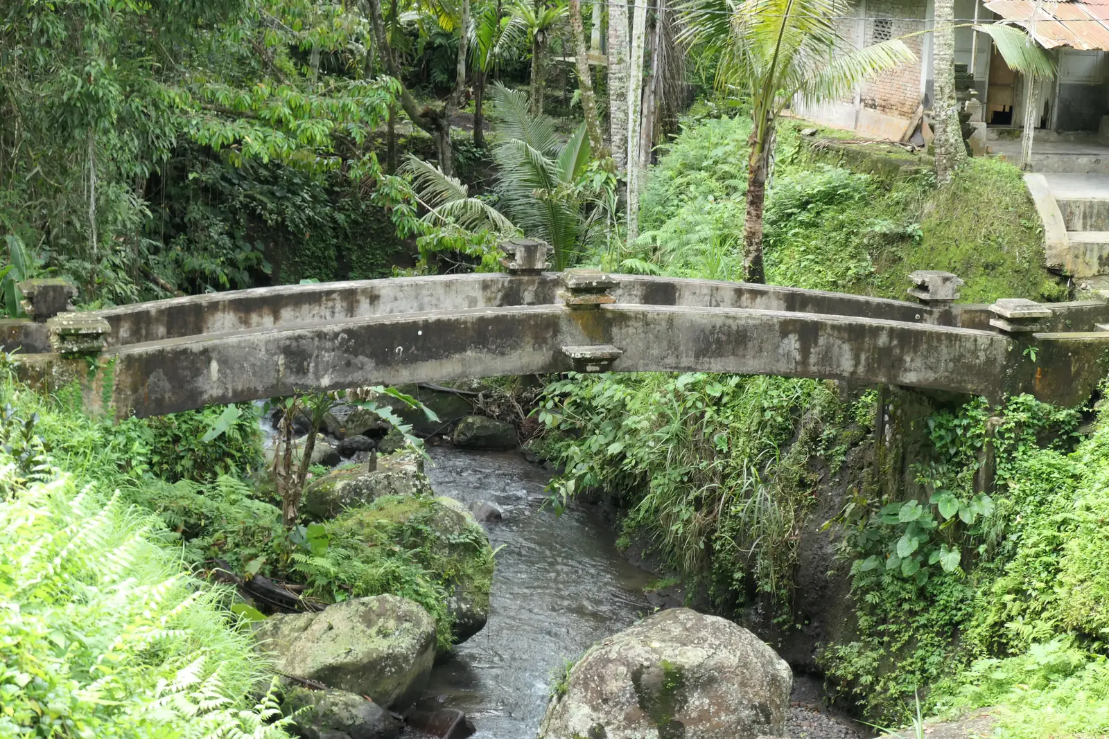 Jembatan beton kecil melintasi Sungai Pakerisan di antara pepohonan dan batu sungai pada kawasan Gunung Kawi